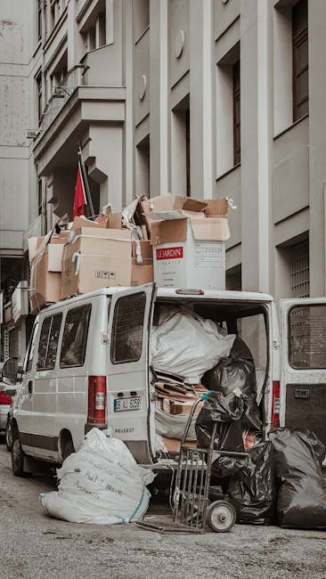 A white panel van parked on a city street in front of a multi-storey building with a grey facade and numerous windows. The van's rear doors are open, revealing a large pile of packed waste, including cardboard boxes, plastic bags, and wrapping materials, some of which are spilling out onto the pavement. The cardboard boxes vary in size and are stacked loosely, with some labeled and others plain. A small hand trolley is positioned beside the van, loaded with black garbage bags. The scene reflects the process of loading bulky waste in preparation for removal, consistent with home relocation or waste clearance services offered by Man with Van Wapping, situated on a typically urban environment suitable for removals and furniture transport activities.