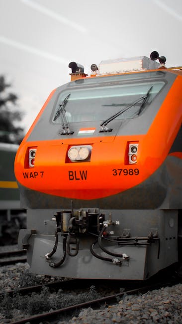 A close-up of the front section of an orange and gray passenger train positioned on railway tracks during daylight hours. The train's windshield features two windshield wipers, and there are two small lights above a central headlight on the red front panel. The train's identification numbers, 'WAP 7,' 'BLW,' and '37989,' are visible on the front. In the background, blurred electrical wires and tree silhouettes can be seen, with natural lighting casting a neutral tone over the scene. This image, associated with house removals and moving logistics, captures the loading or transportation process of large items or equipment, possibly during a home relocation or furniture transport operation, as facilitated by [COMPANY_NAME] or similar removal service providers.