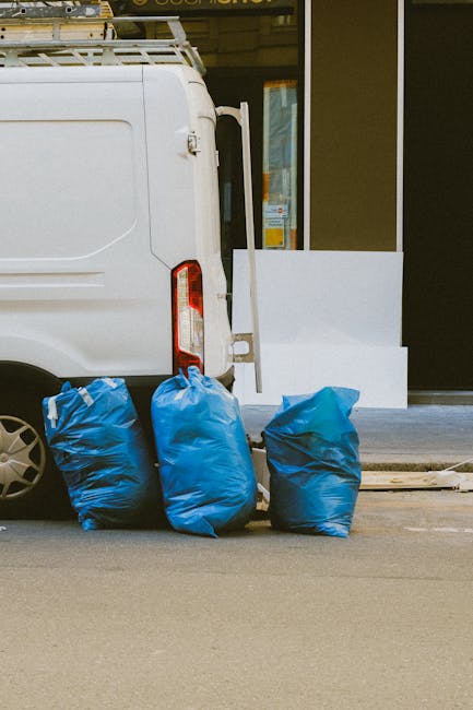 A white panel van parked on a city street in front of a multi-storey building with a grey facade and numerous windows. The van's rear doors are open, revealing a large pile of packed waste, including cardboard boxes, plastic bags, and wrapping materials, some of which are spilling out onto the pavement. The cardboard boxes vary in size and are stacked loosely, with some labeled and others plain. A small hand trolley is positioned beside the van, loaded with black garbage bags. The scene reflects the process of loading bulky waste in preparation for removal, consistent with home relocation or waste clearance services offered by Man with Van Wapping, situated on a typically urban environment suitable for removals and furniture transport activities.