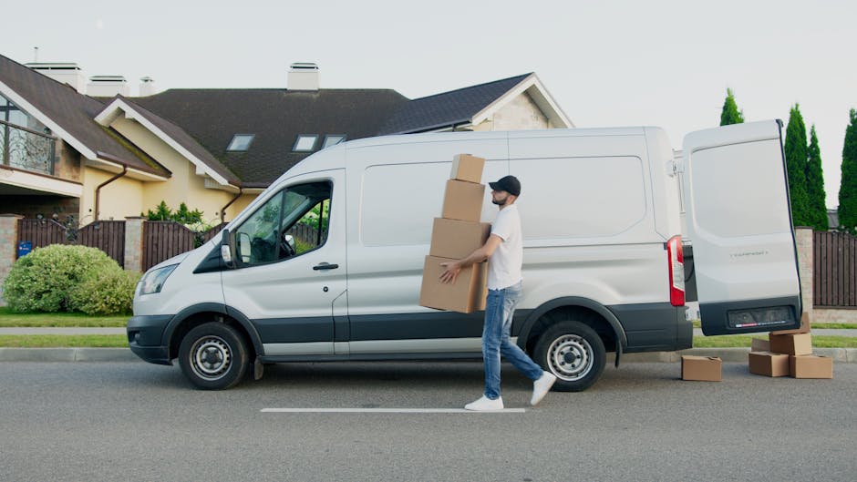 A man wearing a white t-shirt and a black cap is loading a cardboard box into the back of a moving van parked on a driveway, with several other cardboard boxes already stacked inside the vehicle. The 
