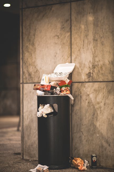 Photograph of a tall, black cylindrical trash bin situated against a beige, tiled wall inside a building, with its white plastic lid open and overflowing with various food packaging and discarded items including wrappers and containers. Nearby on the floor, there are crumpled paper and other small rubbish items. To the right of the trash bin, there is a small canned beverage and a crumpled orange paper bag. The scene is lit with indoor ambient lighting, and the overall setting appears to be a public space or hallway. This image relates to waste disposal as part of a house or office move, relevant to the context of packing and clearing clutter during a relocation process, which is supported indirectly by the expertise of Man with Van Wapping.