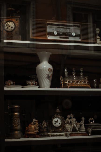A close-up view of a glass-fronted wooden cabinet containing various antique and vintage household items, including a white ceramic vase with floral decoration on the middle shelf, ornate glass perfume bottles with stoppers on the top shelf, and a brass and ceramic clock along with small decorative figurines and jewelry boxes on the bottom shelf. The objects are arranged in a neat display, with soft lighting highlighting their intricate details. The scene appears to be inside a home, possibly a living room or a storage area, where Man with Van Wapping is involved in house removals. The careful packing of fragile items such as glass bottles and delicate ornaments suggests a furniture transport or packing process as part of home relocation efforts.