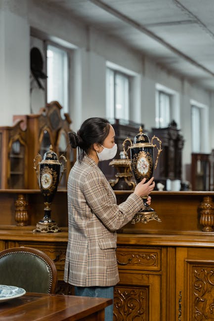 A woman wearing a protective face mask and a checked blazer is examining and handling two large, ornate antique urns with intricate gold and black decorative details. She is standing inside a spacious, well-lit room with large windows and a high ceiling, surrounded by other antique furniture and wooden cabinetry. The urns are placed on a long, polished wooden surface, possibly a sideboard or table, which is part of a collection of vintage decorative items. The setting appears to be an antique shop or a storage area for relocation purposes, where careful handling and packing of valuable antiques are important. The lighting is natural, coming from the windows, highlighting the details of the antiques and the woman's focused activity, consistent with professional house removals or relocation services like those provided by Man with Van Wapping.