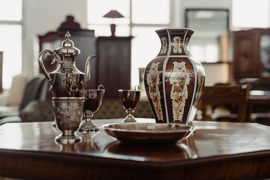 A close-up view of a glass-fronted wooden cabinet containing various antique and vintage household items, including a white ceramic vase with floral decoration on the middle shelf, ornate glass perfume bottles with stoppers on the top shelf, and a brass and ceramic clock along with small decorative figurines and jewelry boxes on the bottom shelf. The objects are arranged in a neat display, with soft lighting highlighting their intricate details. The scene appears to be inside a home, possibly a living room or a storage area, where Man with Van Wapping is involved in house removals. The careful packing of fragile items such as glass bottles and delicate ornaments suggests a furniture transport or packing process as part of home relocation efforts.