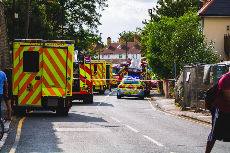 A street scene in Wapping showing multiple emergency service vehicles, including a yellow and red London ambulance and several fire engines, parked along the roadway with their blue lights flashing. The vehicles are positioned in a line blocking part of the street, which is cordoned off with red and white tape. People are walking nearby, some observing the scene. The surroundings include residential houses with brick exteriors, trees, and fencing along the pavement. The emergency services staff are visible attending to the situation, and the environment appears bright and outdoors. This scene reflects a response by emergency personnel, possibly involving the safe evacuation or rescue operation at a residential property, which aligns with the context of house removals, packing, and moving logistics managed by professionals such as Man with Van Wapping.