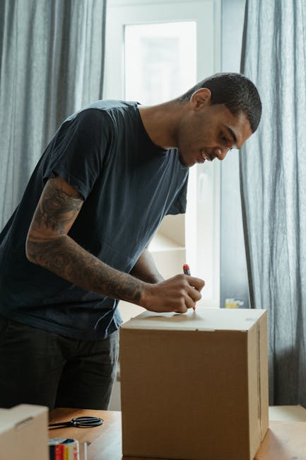A man with short dark hair and a tattooed arm is inside a well-lit room, standing next to a wooden box while using a red marker to label or seal it during a home relocation process. Behind him, a large window with partly open light blue curtains allows natural daylight to illuminate the space. On the table in front of him, there are various packing materials, including a pair of scissors and pieces of tape. The scene depicts the packing and moving stage of a house removal, with the man preparing items for transport. This activity is part of the logistics involved in furniture transport and packing for a move, with the focus on careful labeling and securing of belongings before loading into a van. The setting suggests a professional approach to same-day emergency moves in Wapping, with [COMPANY_NAME], Man with Van Wapping, providing reliable removals services.
