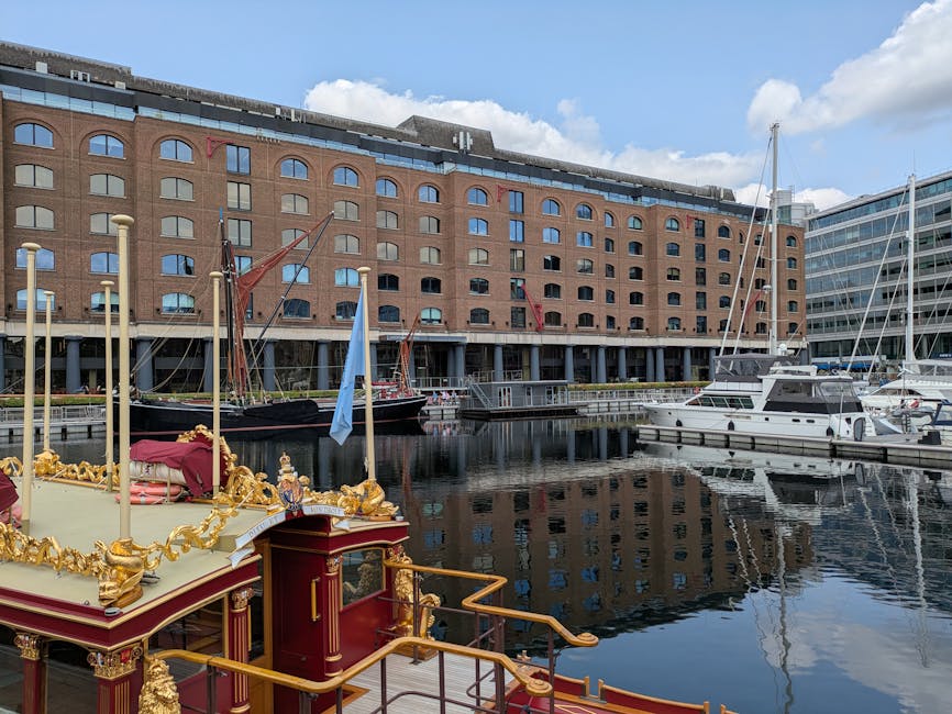 A waterfront scene featuring a variety of boats and yachts docked along the quay, with a prominent large brick building in the background. The boats include a decorative boat with ornate gold embellishments and red fabric on its upper deck, and several modern motor yachts with white hulls and masts. The ships are secured to the docks, and some are covered with protective tarps or fabrics. The setting suggests a busy marina environment, often involved in home relocation or furniture transport activities. The sky is partly cloudy, and the reflection of the boats and the building can be seen on the calm water. This image is associated with moving services such as those provided by Man with Van Wapping, illustrating the logistical aspects of loading or unloading vessels as part of a relocation process near Wapping.