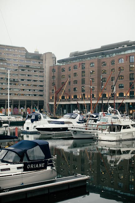 A row of moored sailboats and yachts situated along a canal or river, with colorful British flags and bunting attached to their masts. The boats are anchored close to a pavement, where a large green tree provides partial shade. Behind the waterfront, there is a multi-story brick building featuring arched windows, with modern glass skyscrapers rising in the background, highlighting a contrast between traditional and contemporary architecture. The scene is well-lit, suggesting daytime with clear weather. This setting illustrates the typical environment involved in house removals or furniture transport near waterfront locations such as Wapping, and may be relevant to services like packing, loading, or moving logistics offered by companies like Man with Van Wapping.