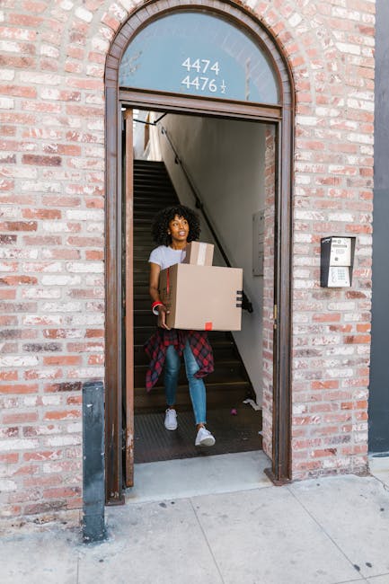 A young woman with curly hair is emerging from the entrance of a brick building, carrying a cardboard box and a smaller package, both secured with packing tape. She is dressed in casual clothes, including a white t-shirt, blue jeans, and white sneakers, with a red and black checked shirt tied around her waist. The entrance features an arched window above the door displaying the numbers '4474' and '4476,' and a modern intercom system is mounted on the right side of the doorway. Inside, a staircase with dark steps and a metal handrail is visible, indicating an interior space used for residential or commercial purposes. The woman appears to be part of a house or office relocation, with a focus on packing, moving, and furniture transport services provided by [COMPANY_NAME], as part of their house removal and relocation logistics. The scene is set during daytime with natural lighting highlighting the details of the brickwork, packaging, and her movement within the loading area of the property.