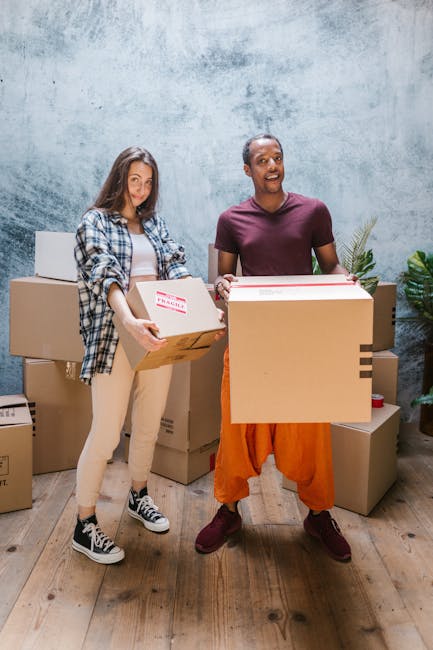 Two individuals, a woman and a man, are standing indoors in front of a plain, textured grey wall with a wooden floor, participating in a home relocation or packing process. The woman, dressed casually in a plaid shirt, light-colored trousers, and black-and-white sneakers, is holding a cardboard box labeled 'Fragile' with both hands, prepared for furniture transport or moving. The man, wearing an orange protective apron over a dark purple T-shirt and maroon shoes, is smiling broadly while carrying a larger cardboard box, likely filled with household items. Several other cardboard boxes, some sealed and others partially open, are stacked around them, indicating an active packing or loading process. Nearby, a portable trolley and packing supplies might be visible, although not clearly detailed. The scene exemplifies organized packing, part of a professional removals service by Man with Van Wapping, supporting efficient local house moves in the Wapping area, consistent with services described for house removals, furniture transport, and packing and moving logistics.