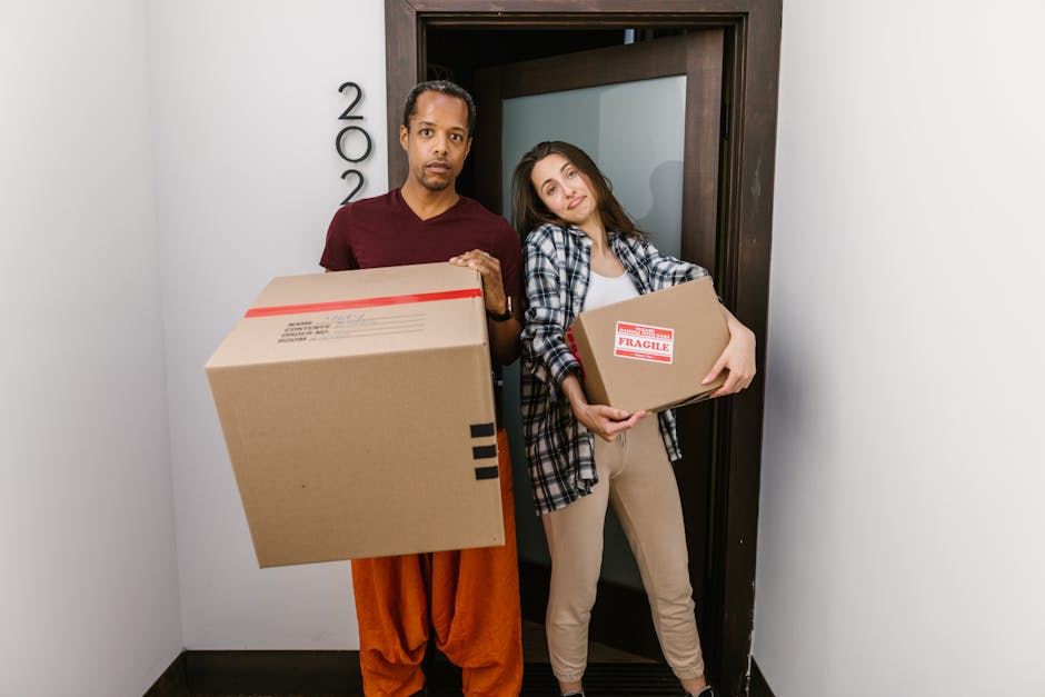 A young woman with curly hair is emerging from the entrance of a brick building, carrying a cardboard box and a smaller package, both secured with packing tape. She is dressed in casual clothes, including a white t-shirt, blue jeans, and white sneakers, with a red and black checked shirt tied around her waist. The entrance features an arched window above the door displaying the numbers '4474' and '4476,' and a modern intercom system is mounted on the right side of the doorway. Inside, a staircase with dark steps and a metal handrail is visible, indicating an interior space used for residential or commercial purposes. The woman appears to be part of a house or office relocation, with a focus on packing, moving, and furniture transport services provided by [COMPANY_NAME], as part of their house removal and relocation logistics. The scene is set during daytime with natural lighting highlighting the details of the brickwork, packaging, and her movement within the loading area of the property.