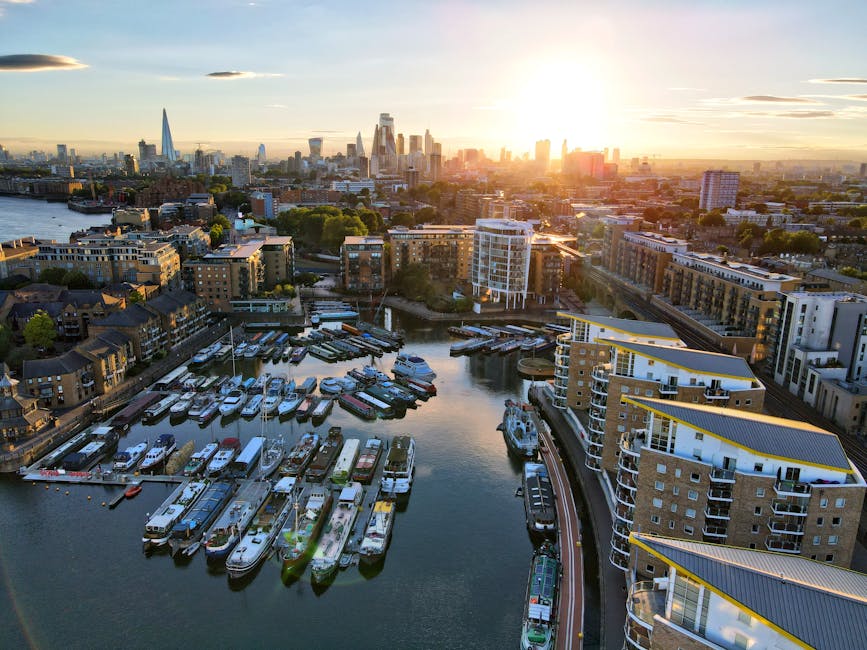 An aerial view of Tower Hamlets in Wapping during sunset, showing a mix of modern and historic buildings, marina filled with various boats and yachts, and waterways reflecting the golden light. The scene includes residential apartments atop a residential complex on the right, with visible balconies and exterior corridors, and boats secured along the quayside. In the distance, the skyline features prominent London skyscrapers, including The Shard, with the setting sun casting a warm glow across the cityscape. This urban environment illustrates the type of area where house relocations and furniture transport services by Man with Van Wapping are often required, especially when securing permits for moving within or through Wapping, as part of efficient packing and loading processes during home relocation projects.