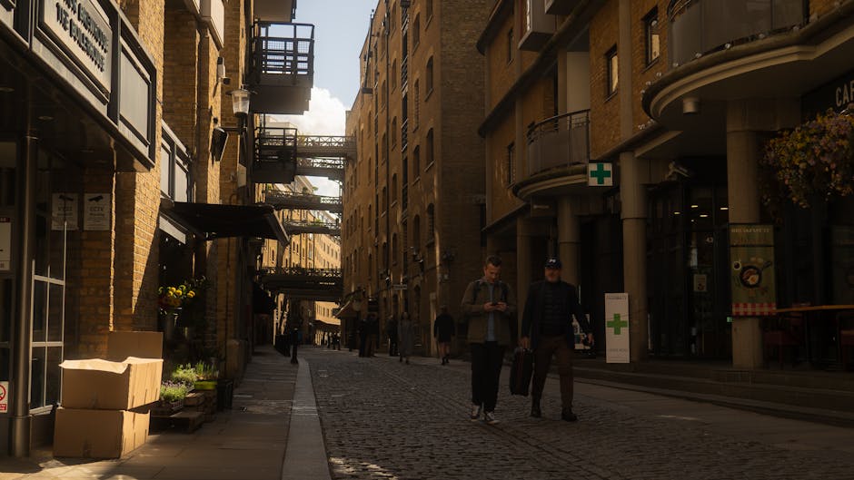 The image shows a cobbled street scene in Wapping with several multi-storey brick buildings on either side, featuring external fire escape staircases and balconies. In the foreground, cardboard boxes and plant pots are placed on the pavement near the entrance of a building, indicating packing or unpacking activities. To the right, a man wearing a dark jacket and hat is walking alongside another man dressed casually, both carrying luggage or belongings, suggesting a home relocation or moving process supported by Man with Van Wapping. Behind them, a few pedestrians are visible, some also carrying items, while others are strolling along the street. The street is illuminated by natural daylight, with shadows cast by the buildings, and a shop with a green cross sign indicating a pharmacy can be seen on the right. The overall setting captures the typical environment of a residential area involved in furniture transport and packing during a house move, consistent with a professional removals service at work in an urban context.