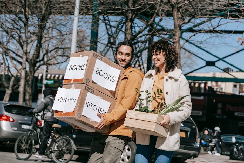 A man and a woman are outdoors, engaged in house moving activities, with the man holding two large cardboard boxes labeled 'BOOKS' and 'KITCHEN' for home relocation, and the woman carrying a wooden tray with potted plants. The boxes are secured with packing tape and have printed labels, indicating careful packing and organisation typical of a furniture transport or packing and moving process. They are positioned on a pavement near a street with parked cars, bicycles, and leafless trees under a clear sky, suggesting a daytime move. The background includes a blue building or scaffolding, and the scene captures a typical step in a home relocation involving loading or unloading belongings. This image exemplifies the physical aspect of moving services provided by companies such as Man with Van Wapping, emphasizing packing, carrying, and transport logistics in a residential setting.