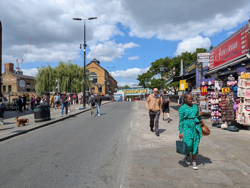 The image shows a cobbled street scene in Wapping with several multi-storey brick buildings on either side, featuring external fire escape staircases and balconies. In the foreground, cardboard boxes and plant pots are placed on the pavement near the entrance of a building, indicating packing or unpacking activities. To the right, a man wearing a dark jacket and hat is walking alongside another man dressed casually, both carrying luggage or belongings, suggesting a home relocation or moving process supported by Man with Van Wapping. Behind them, a few pedestrians are visible, some also carrying items, while others are strolling along the street. The street is illuminated by natural daylight, with shadows cast by the buildings, and a shop with a green cross sign indicating a pharmacy can be seen on the right. The overall setting captures the typical environment of a residential area involved in furniture transport and packing during a house move, consistent with a professional removals service at work in an urban context.